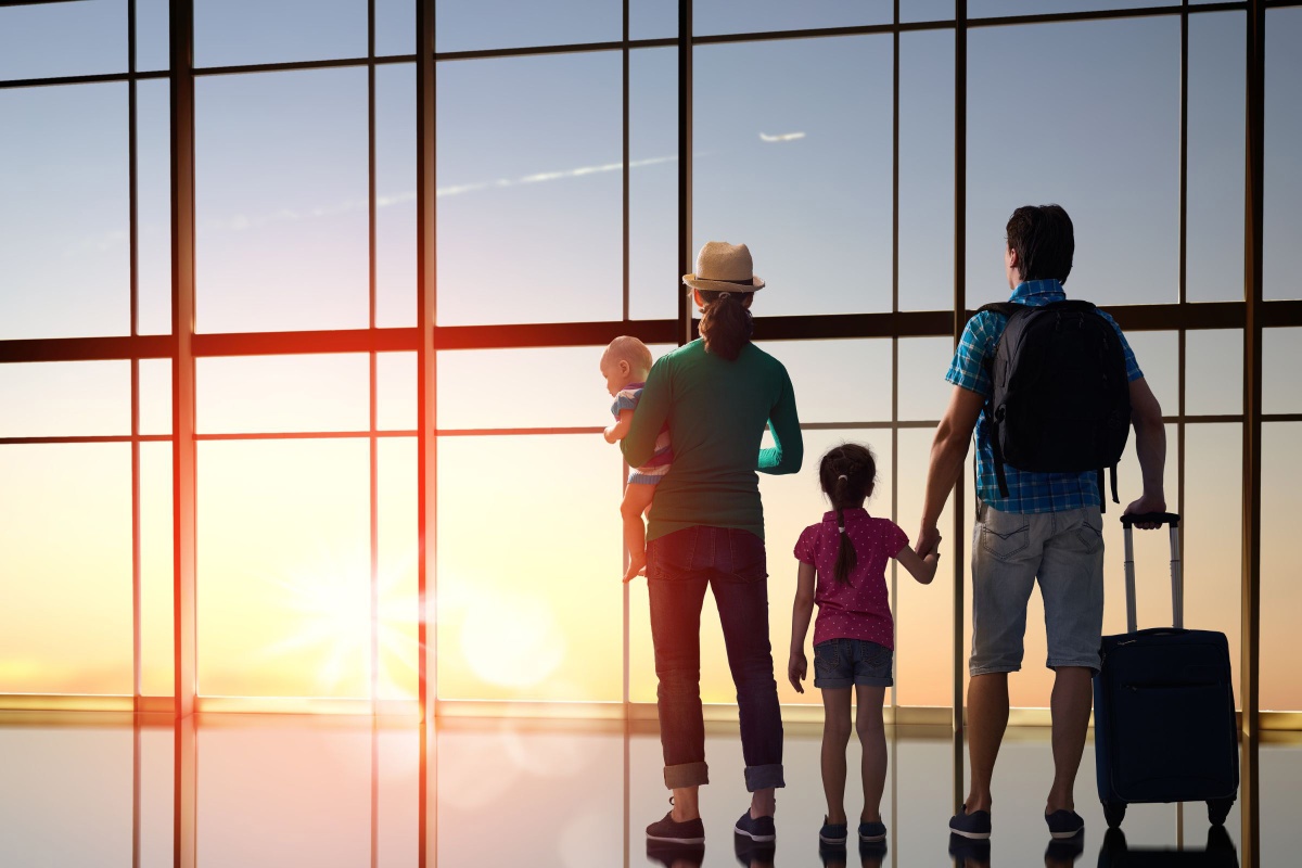 Family with young children eagerly waiting for their flight at the airport. The parents and kids are standing near a large window, gazing at planes taking off and landing. 