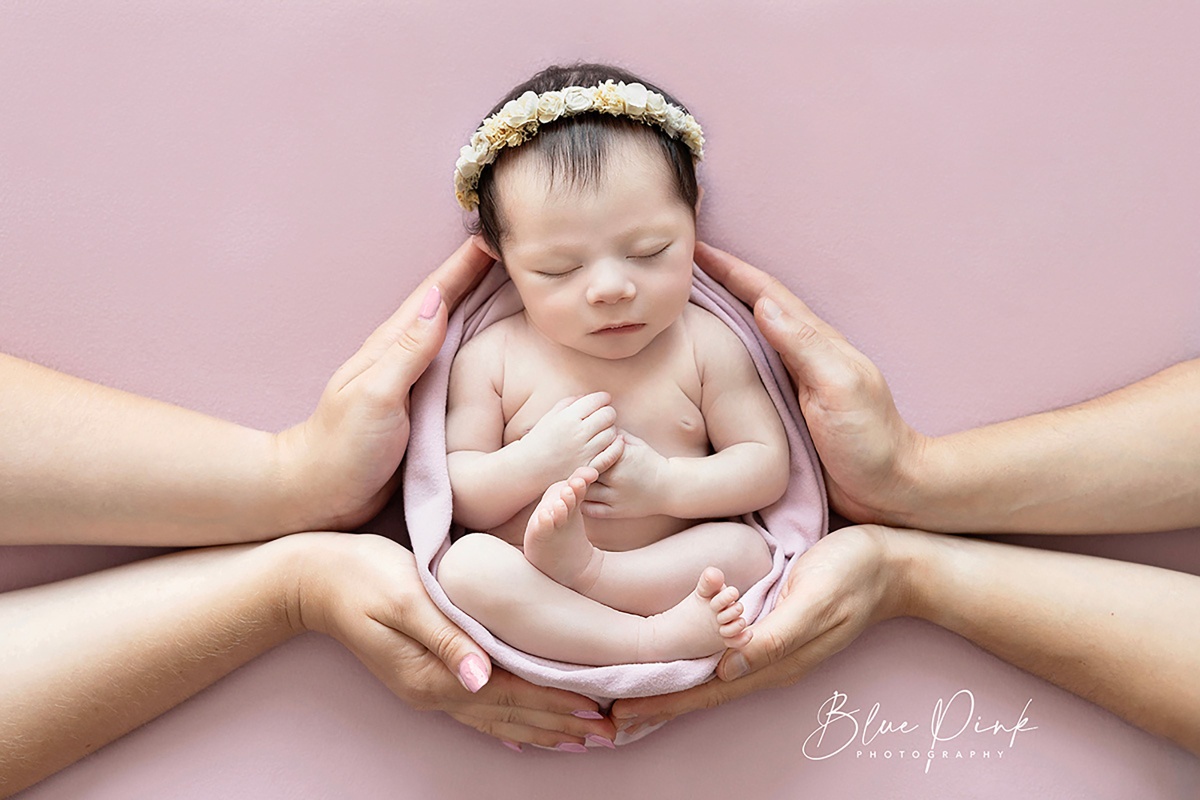 Adorable newborn baby, wrapped in a delicate pale pink elastic wrap, cradled by the supportive hands of loving parents on both sides.