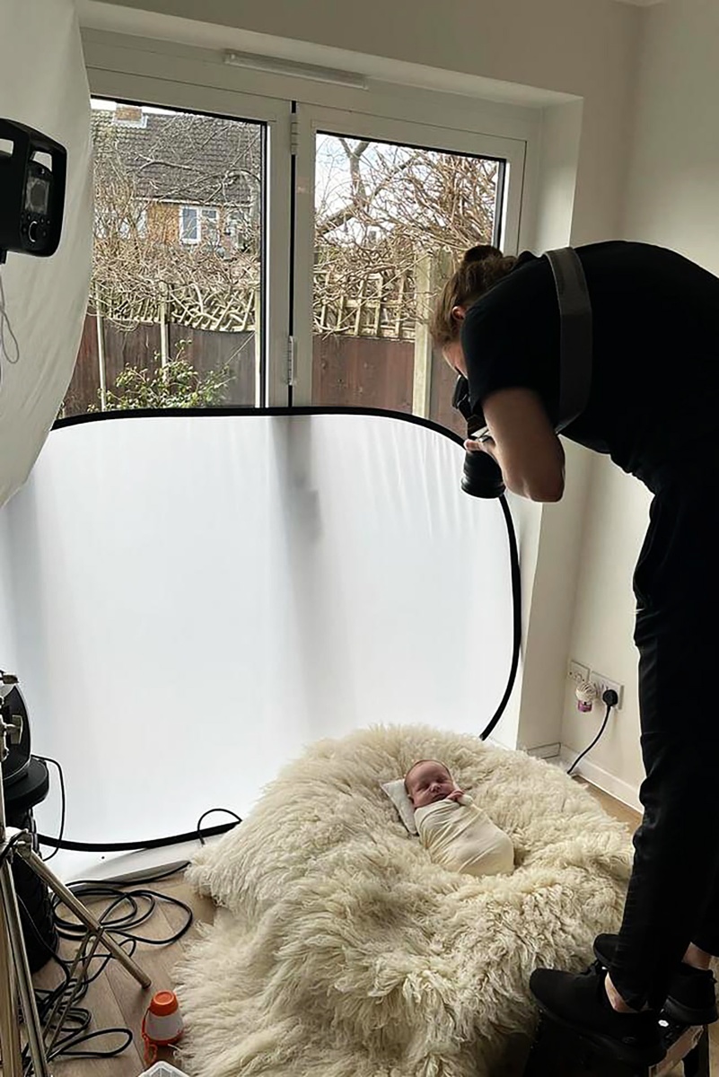A professional newborn photographer capturing images of a gently wrapped newborn lying on natural fur during a behind-the-scenes session.