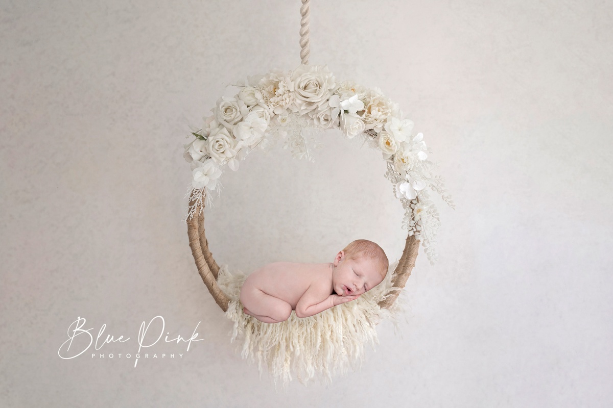 Adorable naked newborn baby comfortably curled on his tummy, resting on a soft white fur surface within a stunning white flower circle in boho style. 