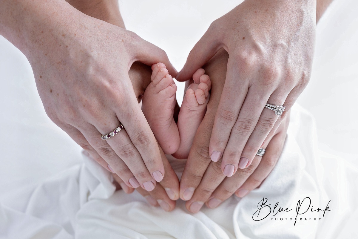 The delicate feet of a newborn are cradled in the loving hands of the parents against a bright white fabric backdrop. 