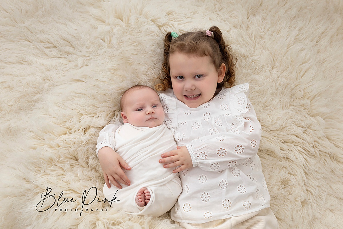 Adorable six-year-old sister lovingly holds her newborn baby brother wrapped in a soft white elastic wrap, both lying on a cream-colored flokati rug, facing the camera.