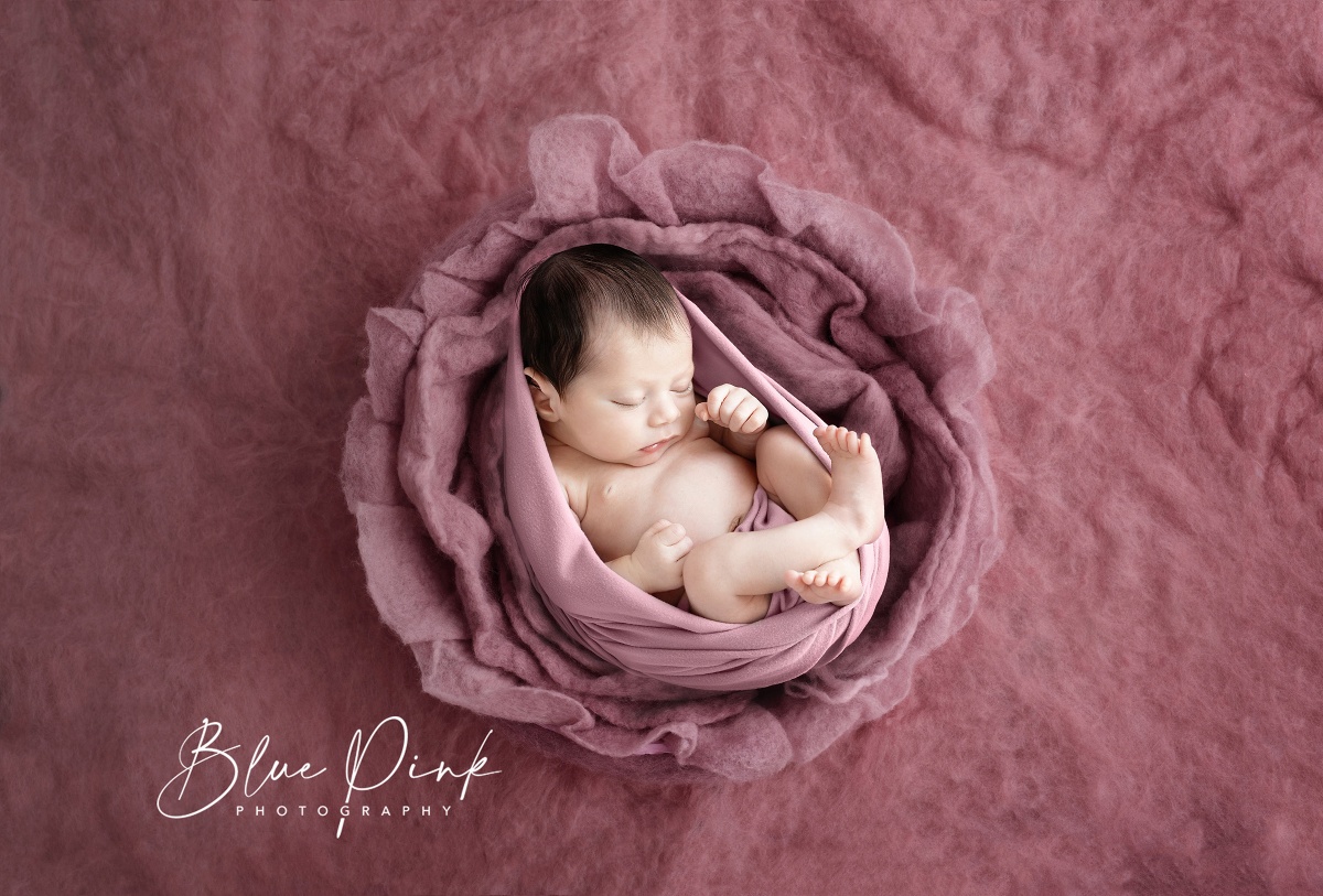 A newborn baby girl peacefully lays on her back in a rose pink wool flower nest, gently wrapped in a matching coloured elastic wrap. 