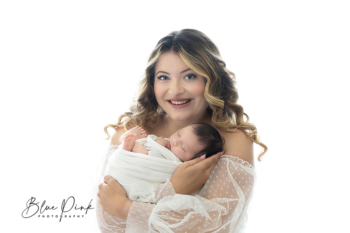 A young mother dressed in a beautiful white classic dress gently holds her newborn baby while facing the camera; the background is white and glowing.