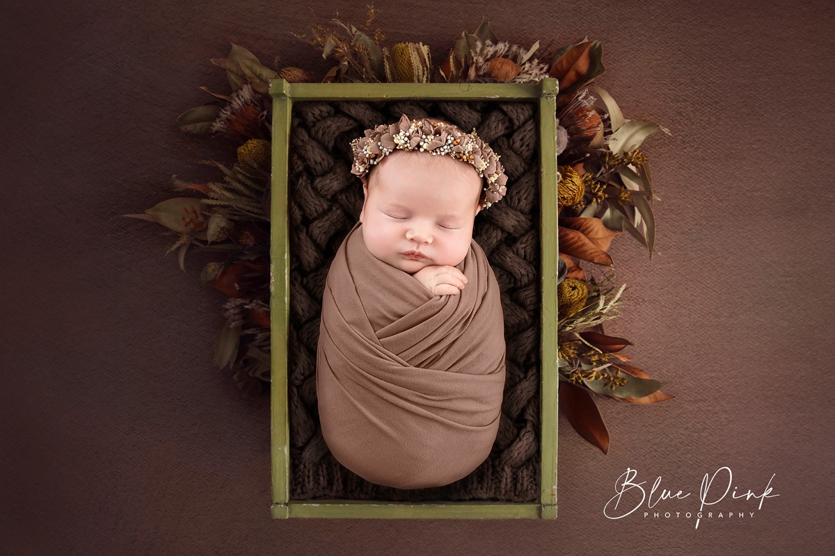 Adorable newborn baby girl, snugly wrapped in a soft brown elastic wrap, lies peacefully in a wooden bed on a soft blanket. A delicate flower halo graces her head, with dry autumn flowers arranged artfully behind the bed.