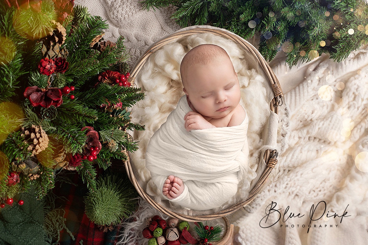 Newborn baby wrapped in white, sleeping in a basket with festive greenery, berries, and cozy textures.