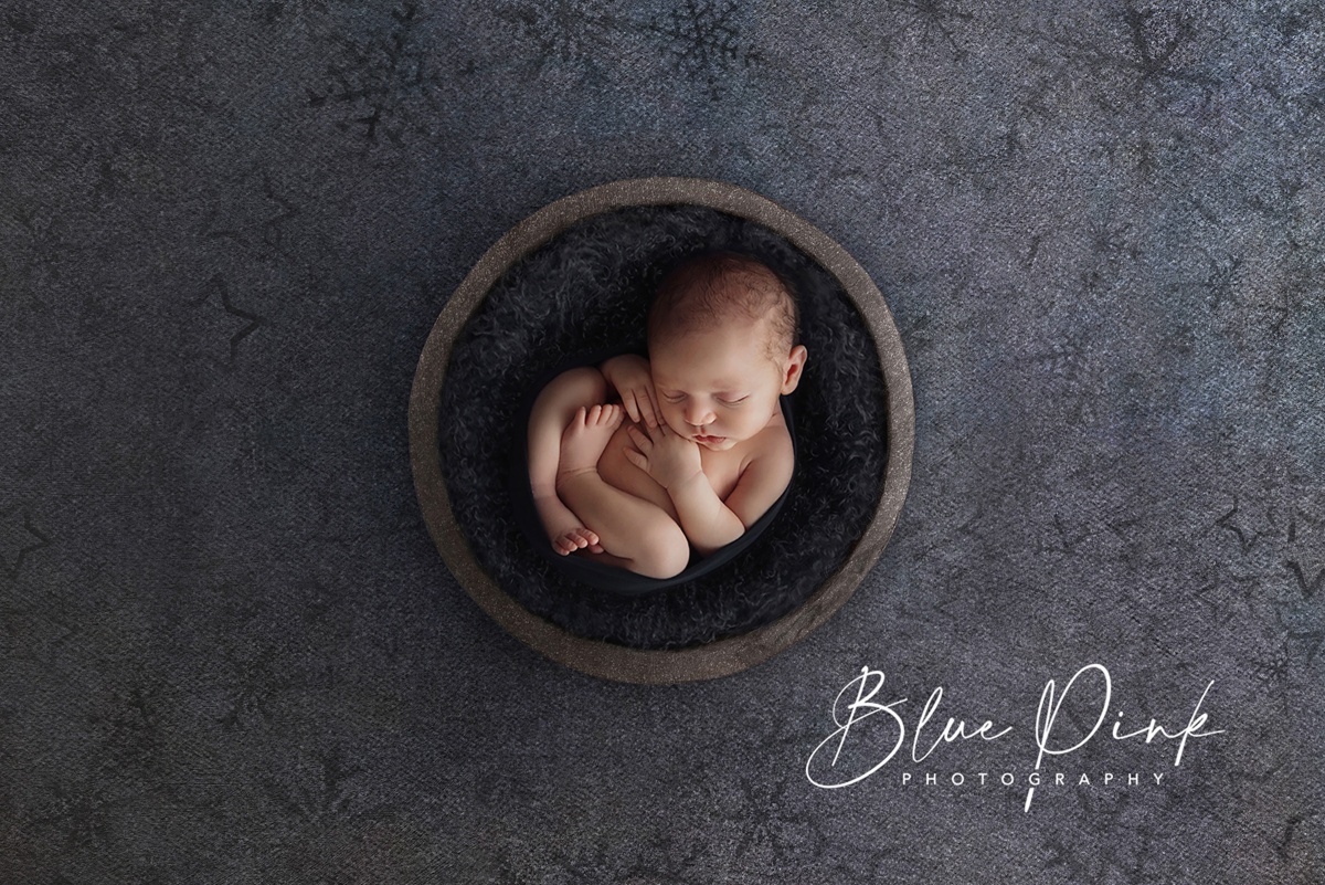 Adorable naked newborn baby delicately posed in a minimalist wooden bowl, set against a deep blue backdrop.