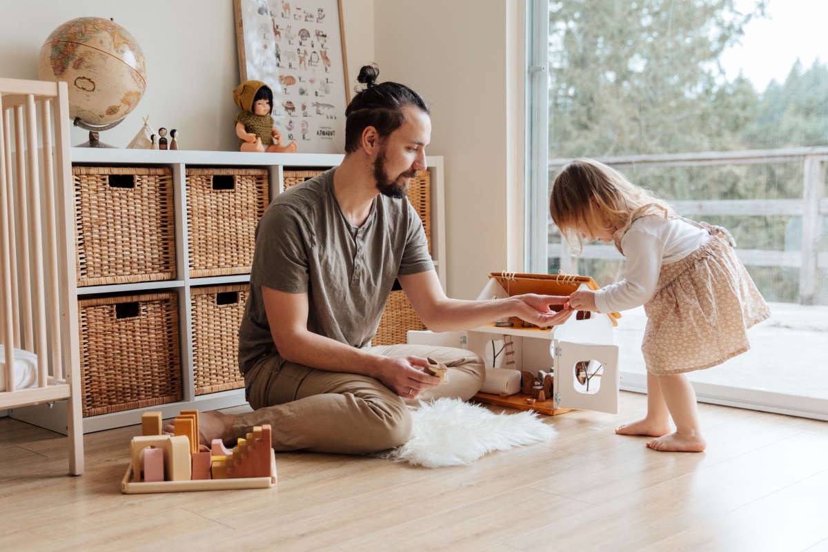 A young dad playing with wooden toys alongside his 3-year-old daughter in a beautifully decorated nursery.