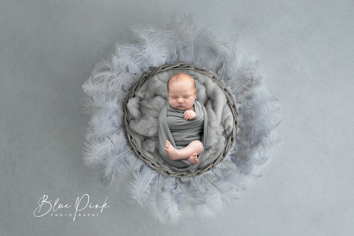 Newborn baby boy peacefully rests inside a wooden basket adorned with a soft grey wool blanket, surrounding the basket are delicate pale blue feathers. 